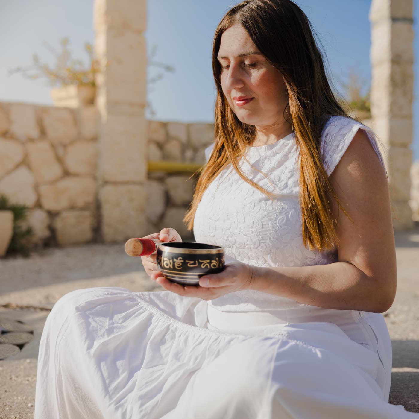 Olga Safa seated outdoors in white, holding a Tibetan singing bowl and its mallet