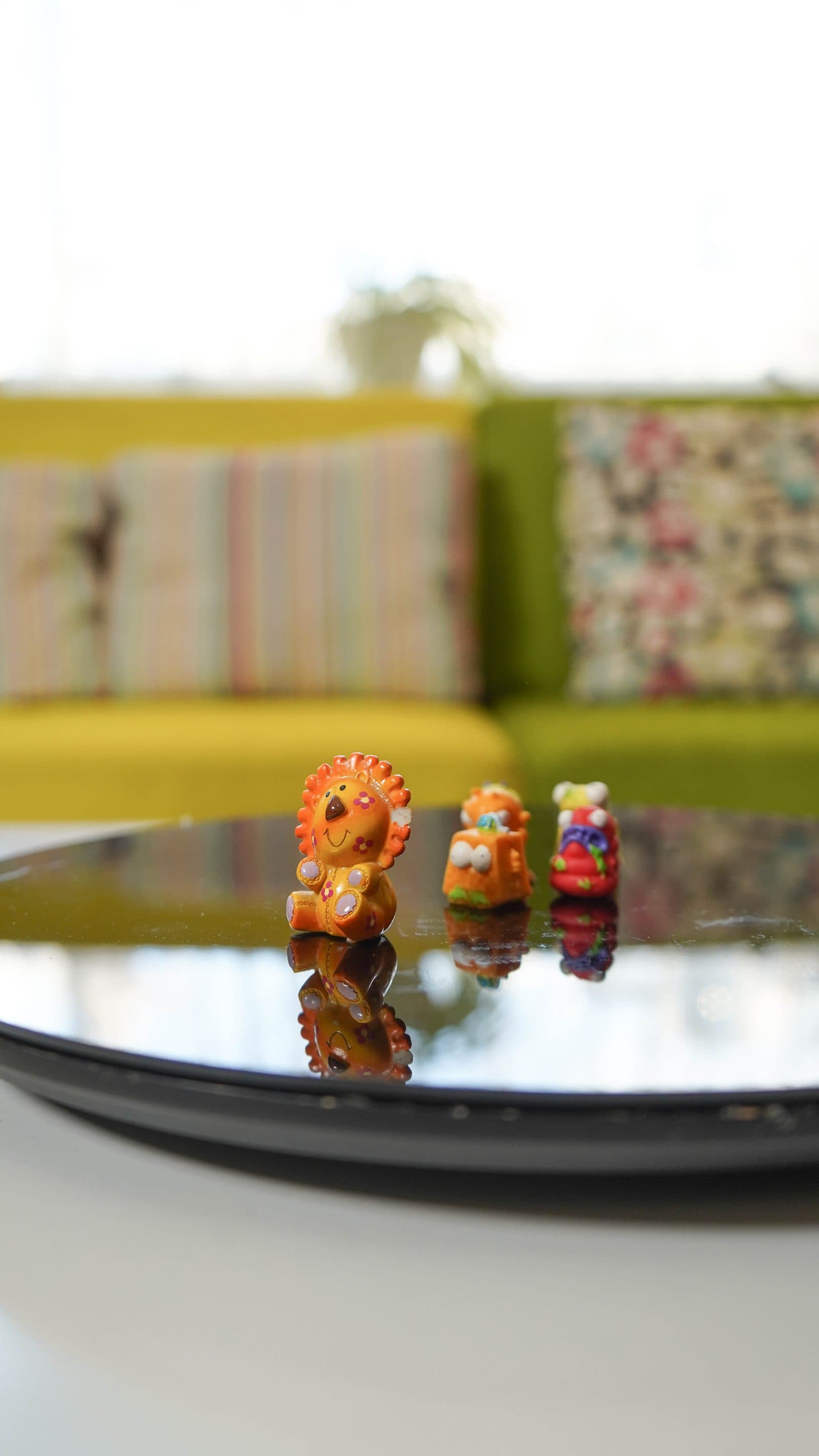 Three small painted figurines standing in a row on a black mirrored plate, a green sofa blurred behind them