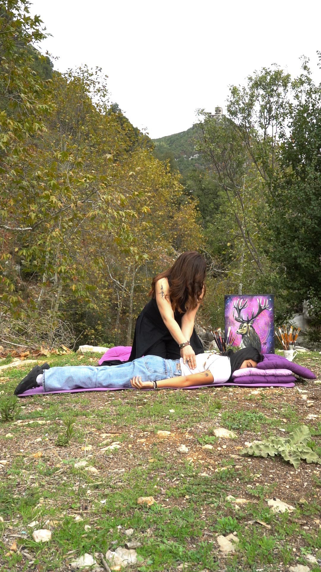 Olga guiding an outdoor breathwork session, hands resting on a participant lying on a mat, wooded hills behind