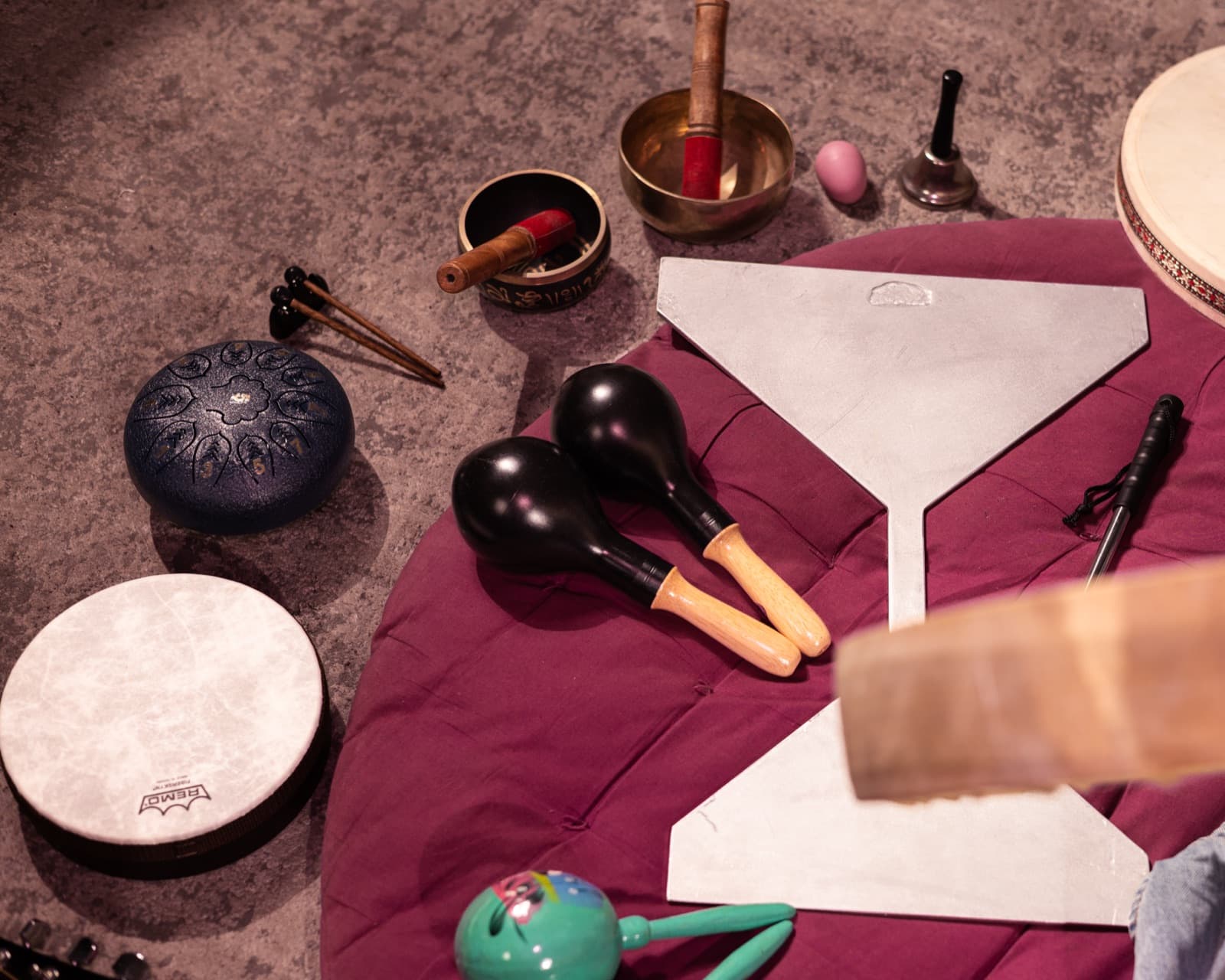 Sound healing instruments laid out on a cushion — singing bowls, a steel tongue drum, frame drums, chime plates, maracas and a bell